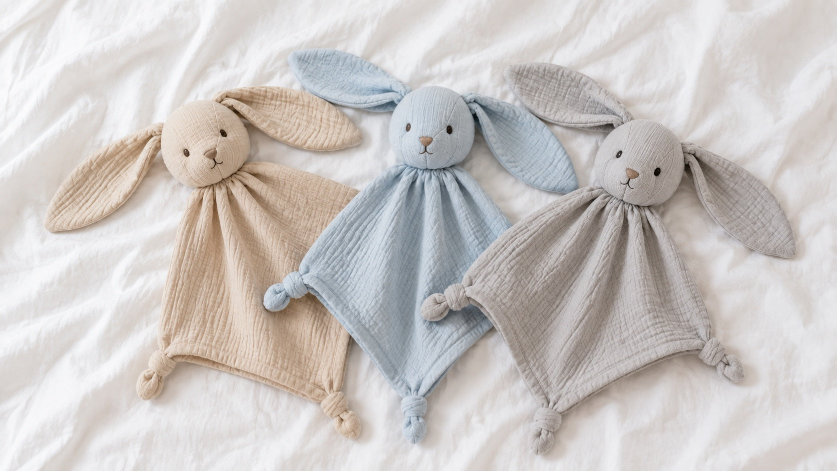 Trois doudous plats lapin, beige, bleu ciel et gris, sur un drap blanc